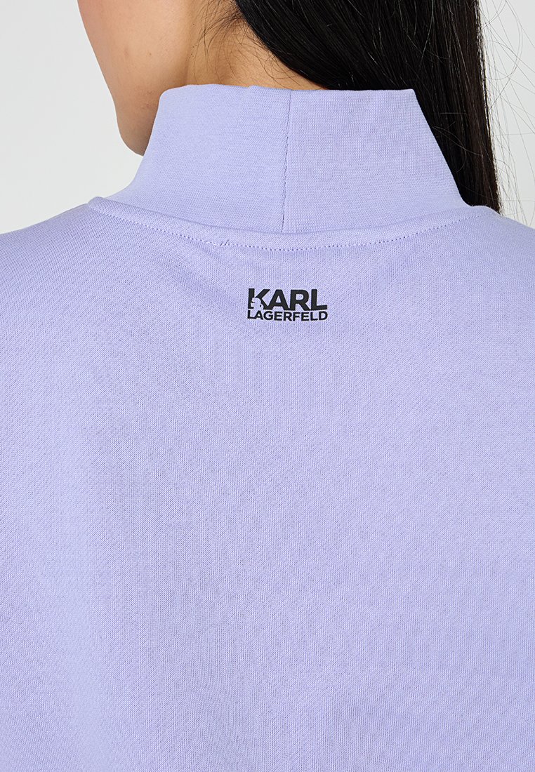 Camisa de manga corta en color lila con cuello alto, que presenta un logo negro de "Karl Lagerfeld" en la parte posterior, textura lisa y un diseño ajustado.