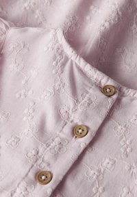Blouse rose clair avec broderies florales, dotée d'un col arrondi et de deux boutons en bois près de l'encolure. Tissu doux et texturé.