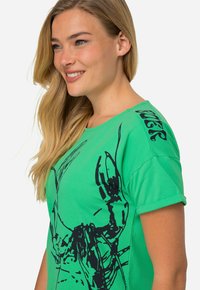 Zelené bavlněné tričko s černým grafickým designem humra na přední straně a logem na rameni. Krátké rukávy, volnější střih.