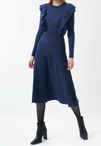 Robe en maille bleu foncé avec détails volants aux épaules, manches longues et jupe plissée mi-longue. Bottines noires complètent le look.
