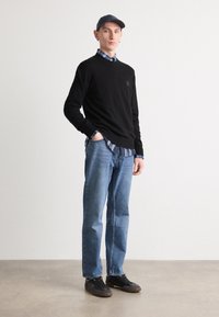 Zwarte trui over een blauw geruit shirt, blauwe jeans en zwarte sneakers. Het model draagt een donkere pet en heeft een slim fit.