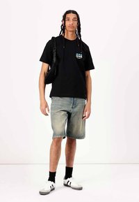 Svart bomulls T-skjorte med grafikk i blått, denim shorts, svarte sokker, og hvite sneakers med svarte striper. Modellen bærer en svart skulderveske.
