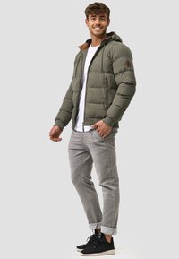 Olijfgroene pufferjack met ritssluiting, capuchon en bruine accenten. Gecombineerd met lichtgrijze broek en zwarte sportieve schoenen.