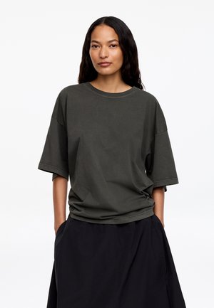 Vrouw met lang donker haar, gekleed in een losvallend donkergrijs T-shirt en een zwarte rok, staand met handen in de zakken tegen een witte achtergrond.