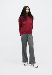 Kvinne med langt mørkt hår iført en burgunder hettegenser, grå joggebukser med sidestriper, og hvite joggesko, stående mot en enkel bakgrunn.