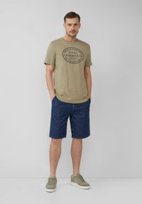 Olchowa koszulka z czarnym okrągłym grafiką, w połączeniu z niebieskimi dżinsowymi szortami. Całość dopełniona szarymi sneakersami, tworząc swobodny wygląd.