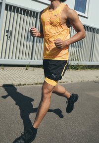 Pánský běžecký outfit skládající se z jasně žlutého a červeného pruhovaného tílka a černých šortek s žlutými akcenty. Doplněno černými sportovními botami.