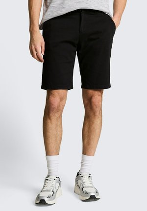Man draagt zwarte knielange shorts, witte sokken en witte sneakers, staat met één hand in de zak tegen een effen achtergrond.