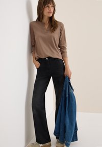 Haut marron à manches longues et jeans noirs. Le mannequin tient une veste en denim bleue. Les baskets sont de couleur marron clair avec des accents blancs. Texture des vêtements lisse.