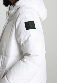 Giacca a vento bianca con cappuccio e cordini elasticizzati, con una toppa quadrata nera sulla manica con la scritta "Calvin Klein" in rilievo.