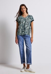 Grünes und schwarzes Schlangenprint-Shirt mit kurzen Ärmeln und V-Ausschnitt, kombiniert mit hellblauen, taillierten Jeans und weißen Sneakers. Flache Textur, lässiges Design.