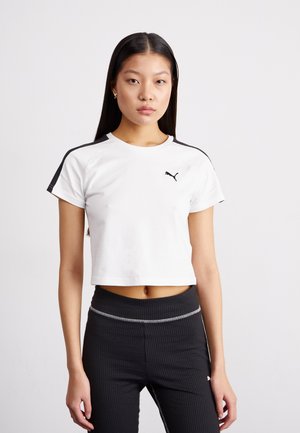 T-shirt blanc cropped à manches courtes, accents noirs sur les épaules et un petit logo Puma noir sur la poitrine. Porté avec des leggings noirs texturés.