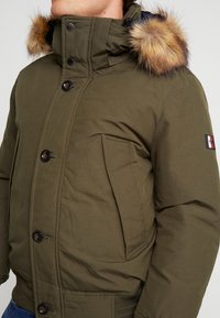 Homme portant une veste d'hiver vert olive avec une capuche bordée de fourrure et une fermeture à boutons à l'avant, montrant le torse et le bras supérieurs.