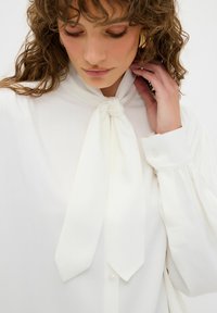 Blouse blanche avec un nœud papillon attaché au col, dotée de longues manches bouffantes et de poignets boutonnés. Texture lisse avec un éclat subtil.
