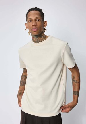 Homme avec des tatouages sur le cou et les bras, portant une chemise unie à manches courtes beige clair et un pantalon foncé sur un fond uni.