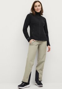 Zwarte fleecejack met hoge kraag, twee zijzakken met ritssluiting; lichtbeige broek met donkere accenten; zwarte schoenen met dikke zolen.