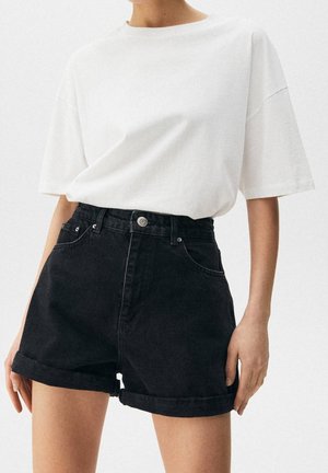 Person wearing a loose white t-shirt tucked into high-waisted black denim shorts with rolled hems, standing against a plain background.