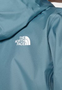 Teal huva jacka gjord av slät tyg, med en framträdande vit logga på vänster sida. Sömmar längs kanterna.