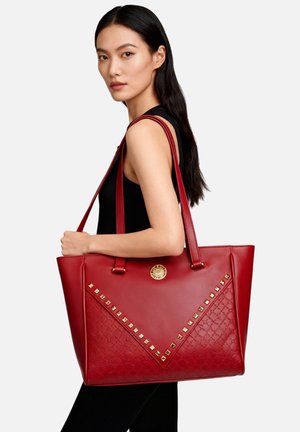 Borsa tote rossa in pelle con design strutturato, caratterizzata da accessori dorati, borchie a forma di piramide e motivo testurizzato su un lato.