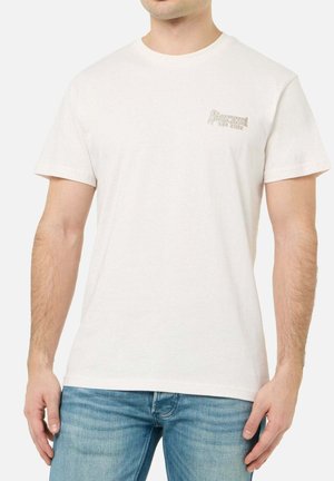 Mann trägt ein schlichtes cremeweißes Kurzarm-T-Shirt mit kleinem "Boxeur Des Rues"-Logo auf der linken Brust, kombiniert mit blauen Jeans.