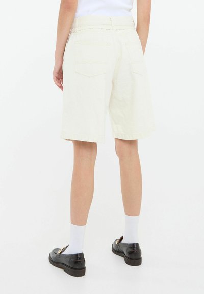 Pantalones cortos de mezclilla color beige claro con un ajuste relajado, que presentan un dobladillo deshilachado y dos bolsillos traseros. Combinados con zapatos negros y calcetines blancos.