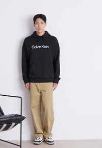 Black hooded sweatshirt with white "Calvin Klein" logo, paired with beige wide-leg pants and black and white sneakers, standing against a white wall.