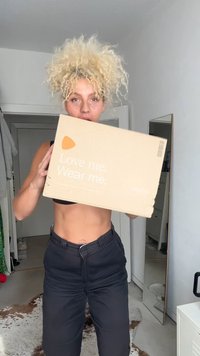 Pappkarton, hellbraun, mit weißem Text "Liebe mich. Trage mich." und einer kleinen orangefarbenen Grafik. Von einer Person in schwarzer Kleidung gehalten.