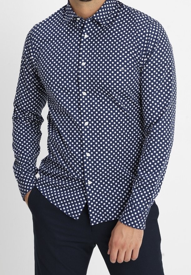 Man in een marineblauwe lange mouw overhemd met witte stippen en donkere broek, hand in de zak op een witte achtergrond.