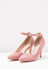 Rosa mocka högklackade skor med spetsig tå, ankelrem och en smal klack. Texturen är slät och mjuk, vilket visar upp en minimalistisk design.