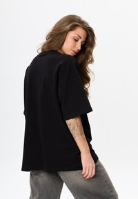 Zwarte oversized t-shirt van zacht materiaal met korte mouwen, voorzien van een ronde halslijn en een relaxte pasvorm. Model draagt grijze, loszittende broek.