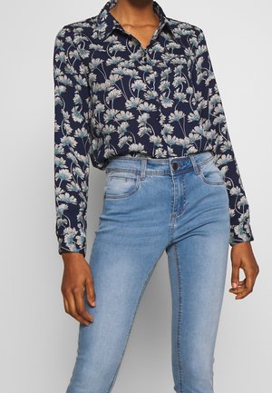 Person wearing a navy floral long-sleeve button-up shirt tucked into light blue high-waisted skinny jeans.
