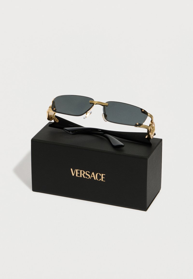 Des lunettes de soleil rectangulaires à monture dorée avec des verres foncés reposant sur une boîte noire Versace avec des lettres dorées.