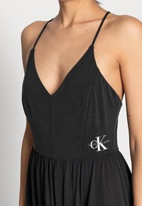 Zwarte jurk met een diepe V-hals, dunne bandjes en een geplooide rok. Heeft een wit Calvin Klein Jeans-logo op het linker taillegebied.