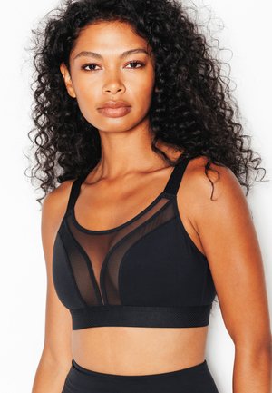 Donna con capelli ricci che indossa un reggiseno sportivo nero con pannelli in rete trasparente, in piedi su uno sfondo bianco.