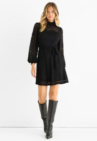 Black lace dress with long sleeves, cinched waist, and skirt. Features a high neckline and a tie belt. Paired with knee-high leather boots.