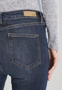 Persona vistiendo jeans de mezclilla azul oscuro con un bolsillo trasero y una etiqueta de cuero marrón claro que dice "edc", vestida con una camiseta de manga larga gris.