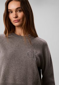 Jeune femme aux cheveux bruns et lisses porte un pull en maille gris avec un discret motif embossé "OK" sur la poitrine, fond gris clair uni.