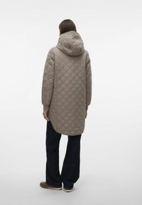 Gequiltede, lange, bruine jas met een capuchon, met een gladde, matte textuur en geribbelde manchetten, gecombineerd met donkergblauwe wijde jeans en casual schoeisel.