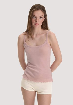 Young woman with long hair wearing a light pink tank top and cream shorts standing against a plain light background.