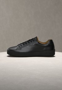 Μαύρο δερμάτινο sneaker χαμηλού προφίλ με μαύρα κορδόνια, ταμπά εσωτερική επένδυση και διακριτικό πλευρικό λογότυπο, εμφανιζόμενο σε ανοιχτή επιφάνεια.