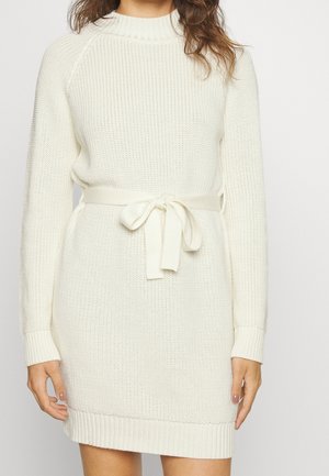 Femme portant une robe-pull blanche en maille à manches longues avec une ceinture nouée à la taille, debout devant un fond uni.