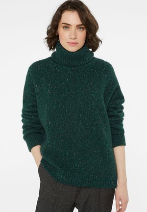 Woman with short brown hair wears dark green turtleneck sweater and gray pants, standing against plain white background.