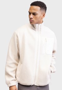 Weiße Fleecejacke mit hohem Kragen, durchgehendem Reißverschluss und Fronttasche. Verfügt über einen strukturierten Stoff und elastische Bündchen für eine enge Passform.