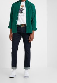 Camisa verde abotonada sobre una camiseta gráfica blanca, vaqueros de mezclilla oscuros con los puños remangados y zapatillas blancas con detalles azules.