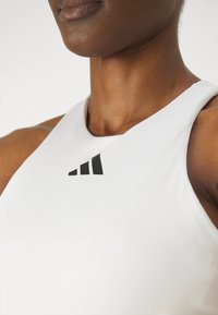Hvit athletic tank topp med høy hals og svart logo på brystet. Stoffet ser jevnt og tettsittende ut.