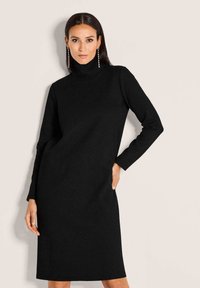Frau mit langen dunklen Haaren, die ein schwarzes langärmliges Rollkragenkleid und hängende Ohrringe trägt, vor einem schlichten beige Hintergrund.