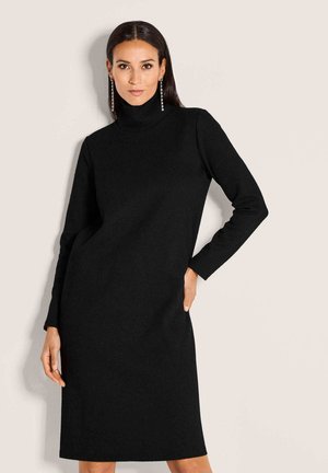 Frau mit langen dunklen Haaren, die ein schwarzes langärmliges Rollkragenkleid und hängende Ohrringe trägt, vor einem schlichten beige Hintergrund.