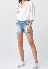 Femme portant une chemise rose clair à boutons, un débardeur blanc, un short en denim bleu délavé et des baskets blanches, debout devant un fond blanc.