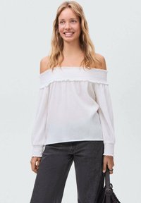 Blusa bianca off-the-shoulder con bordi arruffati e maniche lunghe, realizzata in un tessuto leggero, abbinata a jeans scuri e una borsa nera.