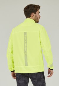 Veste fluorescente jaune avec un col haut, dotée d'un logo réfléchissant et d'accents en mesh sur les manches pour la ventilation et la visibilité.